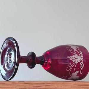May include: A deep red glass goblet lying on its side, featuring a clear stem and a circular base. The bowl of the glass is decorated with an etched white design depicting a cluster of grapes and vine leaves wrapping around the exterior surface.