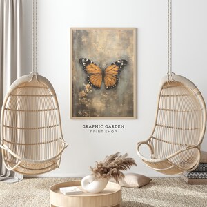 Puede incluir: Una impresi&oacute;n de una mariposa con alas naranjas sobre un fondo texturizado. La impresi&oacute;n est&aacute; enmarcada y colgada en una pared blanca. Dos sillas colgantes de mimbre est&aacute;n en primer plano.