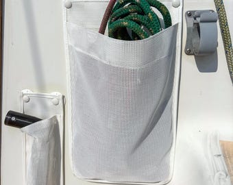Könnte beinhalten: Zwei weiße Netztaschen zur Aufbewahrung, die an einer Bootsfläche befestigt sind. Die größere rechteckige Tasche enthält ein aufgerolltes grünes Seil, während die schmalere Tasche links ein schwarzes zylindrisches Werkzeug enthält. Weiße Druckknöpfe.