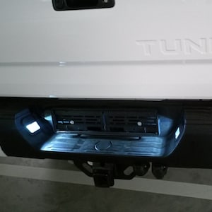 May include: White pickup truck with a black bumper and a license plate holder. The truck has the word "TUNDRA" embossed on the back.