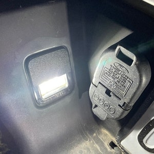 Op de afbeelding: Een close-up van een zwarte plastic aanhangerkoppeling met de tekst "RUNNING LIGHTS", "LEFT STOP TAIL", "BACK UP LIGHTS", "RIGHT STOP TAIL", "TURN SIGNALS", "ELECTRIC BRAKES" en "FORD" erop gedrukt. De connector wordt verlicht door een wit licht.