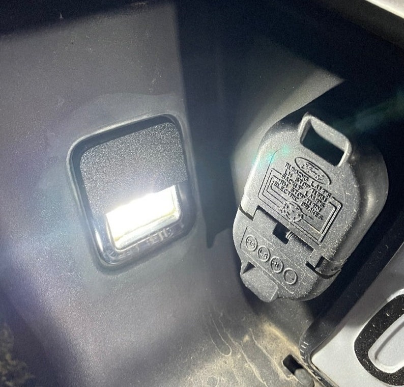 Op de afbeelding: Een close-up van een zwarte plastic aanhangerkoppeling met de tekst "RUNNING LIGHTS", "LEFT STOP TAIL", "BACK UP LIGHTS", "RIGHT STOP TAIL", "TURN SIGNALS", "ELECTRIC BRAKES" en "FORD" erop gedrukt. De connector wordt verlicht door een wit licht.