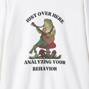 Könnte beinhalten: Weißer Pullover mit einem grünen Frosch, der Gitarre spielt und auf einem Pilz sitzt. Der Text "Just over here analyzing your behavior" ist auf dem Pullover aufgedruckt.
