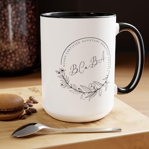 Puede incluir: Taza de cerámica blanca con borde y asa negros. La taza tiene un diseño en blanco y negro con el texto "Board Certified Assistant Behavior Analyst" y las iniciales "BCBA" en una corona de flores.