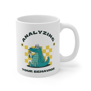 Puede incluir: Taza de cerámica blanca con un dibujo animado de un cocodrilo verde que sostiene un burrito y el texto "ANALYZING YOUR BEHAVIOR" en un círculo negro con estrellas amarillas.