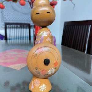 Puede incluir: Muñeca Kokeshi japonesa de madera. La muñeca tiene una cabeza redonda, un cuerpo cilíndrico y una base cónica. Está decorada con diseños florales en naranja, rosa y blanco. La cara de la muñeca tiene los ojos cerrados y una expresión serena. En el fondo, una mesa y un jarrón con bayas rojas.