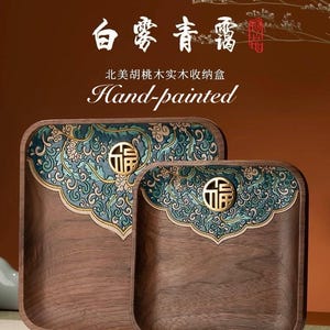 Puede incluir: Dos bandejas cuadradas de madera con un diseño floral pintado a mano en azul y dorado. Las bandejas presentan un carácter chino dorado que se traduce como "buena fortuna". El texto "Hand-painted" está escrito en blanco sobre un fondo marrón. El texto "北美胡桃木实木收纳盒" está escrito en negro sobre un fondo marrón. El texto "白露青霜" está escrito en rojo sobre un fondo marrón.