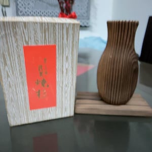 Puede incluir: Un jarrón de madera con ranuras verticales se encuentra sobre una pequeña base de madera. Al lado, una caja con un patrón de vetas de madera y una etiqueta roja con texto dorado. El jarrón y la caja están sobre una superficie reflectante.