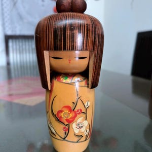 May include: A wooden Kokeshi doll with a brown head and long, vertical hair. The doll's body is tan with a floral design in red, white, and black. The doll has a small, painted face with closed eyes.