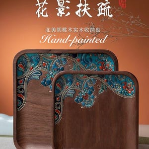 Puede incluir: Dos bandejas cuadradas de servir de madera con diseños florales pintados a mano en azul, verde y rojo. Las bandejas están anidadas y tienen un acabado de madera marrón oscuro. El texto "Hand-painted" está impreso en blanco en la bandeja más grande.