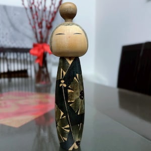 May include: A wooden Kokeshi doll featuring a smooth, rounded head with simple painted facial features and a cylindrical body decorated with a dark green and black floral pattern that wraps around the entire surface of the wood grain.