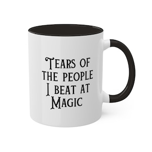May include: White ceramic coffee mug with a black rim and handle. The mug has black text that reads "Tears of the people I beat at magic".