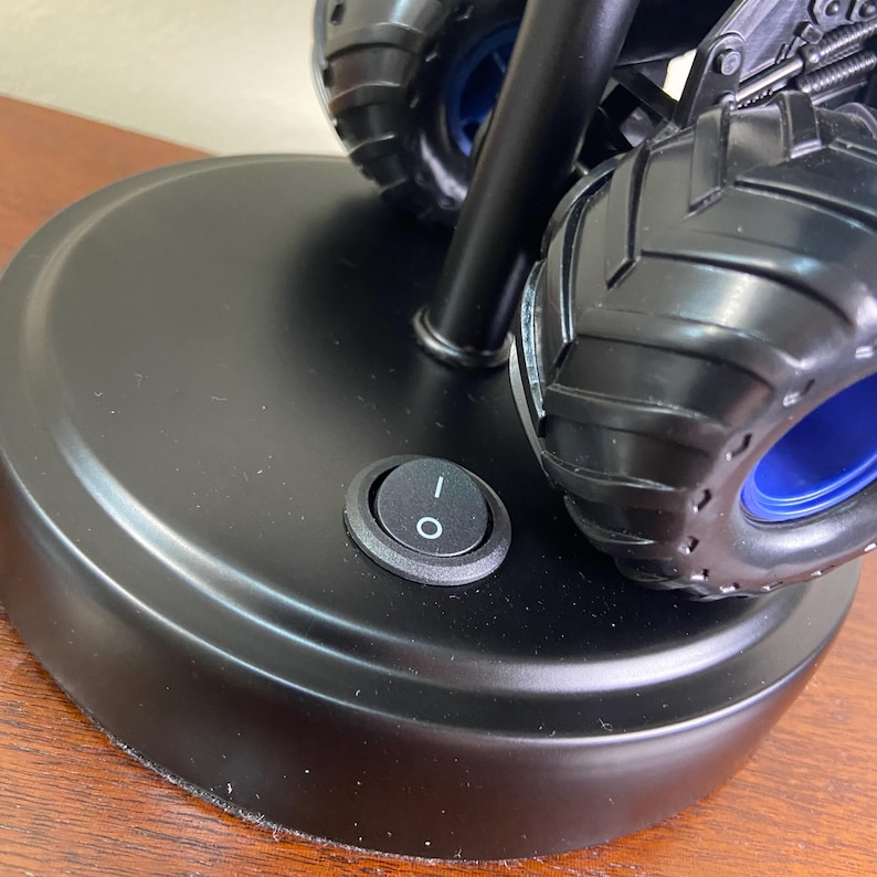 May include: Black monster truck-themed desk lamp with a round base and a toggle switch. The lamp features large, black tires with blue accents. The switch is in the off position.
