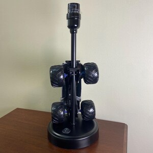 May include: A black monster truck lamp with large, textured tires and a black lamp post. The lamp has a round base with an on/off switch. The tires are black with blue accents.