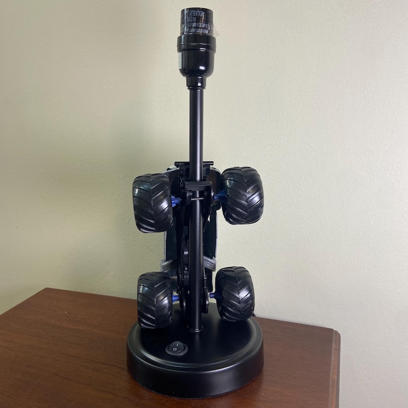 May include: A black monster truck lamp with large, textured tires and a black lamp post. The lamp has a round base with an on/off switch. The tires are black with blue accents.