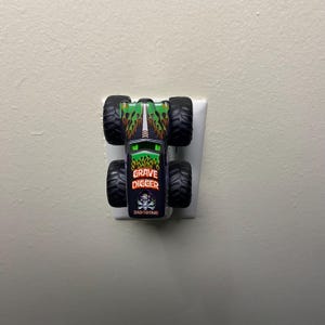 K&ouml;nnte beinhalten: Ein Spielzeug-Monstertruck mit gro&szlig;en schwarzen Reifen und einer schwarzen, gr&uuml;nen und orangefarbenen Karosserie. Der Truck tr&auml;gt die Aufschrift "GRAVE DIGGER" und "BAD BONE". Das Spielzeug ist an einer wei&szlig;en Steckdose angeschlossen.