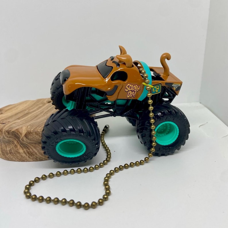 Puede incluir: Un cami&oacute;n monstruo de juguete Scooby Doo marr&oacute;n y negro con neum&aacute;ticos negros grandes y detalles en verde azulado. El juguete tiene las palabras "Scooby Doo!" impresas en el lateral y una cadena dorada.