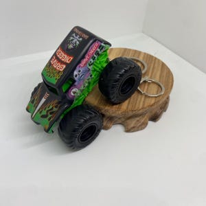 以下が含まれることがあります： ミニチュアのGrave Diggerモンスタートラックのキーホルダー。トラックは黒いボディに緑とオレンジの炎のグラフィックが施され、「Grave Digger」と「Bad Bone」の文字が見えます。タイヤは黒で、キーホルダーは木製の台座に置かれています。