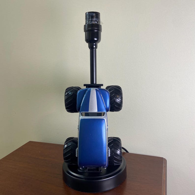 May include: A blue and black monster truck lamp with a black lamp post. The truck has large black tires and a silver stripe down the center. The lamp sits on a black base and is plugged into an electrical outlet.