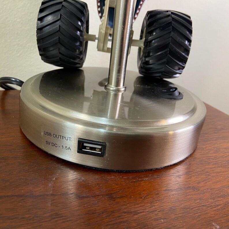 May include: A brushed metal desk lamp with a monster truck design. The lamp features black tires, a USB port, and an on/off switch. The base has the text "USB OUTPUT: 5V DC - 1.5A".