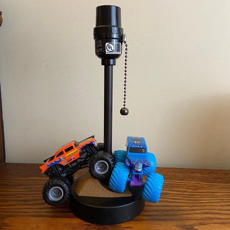 Op de afbeelding: Een zwarte tafellamp met een trekkoord schakelaar. Twee speelgoed monstertrucks staan op de voet van de lamp. Een truck is oranje en zwart met het woord "Avenger" aan de zijkant. De andere truck is blauw en zwart met een schedel en gekruiste botten ontwerp.