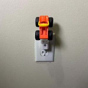 May include: An orange and yellow monster truck night light plugged into a white electrical outlet on a beige wall. The truck has four black tyres and a yellow grille.