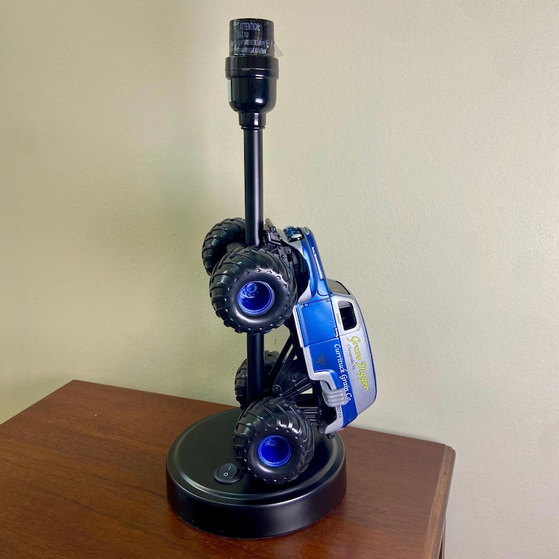 May include: A black table lamp with a monster truck design. The truck is blue and silver with black tires and blue rims. The lamp has a black base and a black pole.
