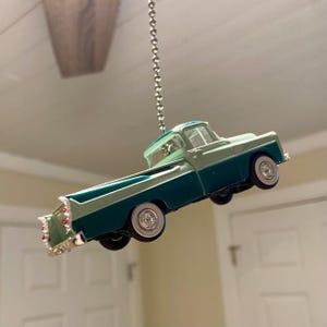 May include: A green and white vintage pickup truck hanging from a silver chain. The truck is a toy model and is suspended in mid-air.