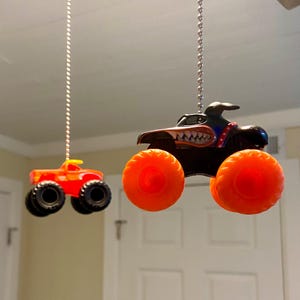 May include: Two toy monster trucks hanging from the ceiling. The larger truck is black with orange wheels and a toothy grin. The smaller truck is red with orange wheels.