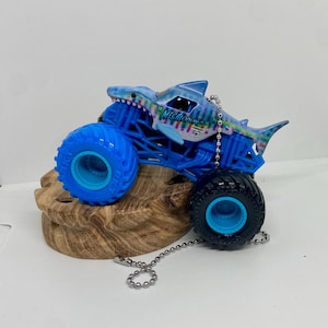 K&ouml;nnte beinhalten: Ein blaues Monstertruck-Spielzeug mit einem Haifisch-Design. Der Truck hat gro&szlig;e blaue Reifen und einen blauen Rahmen. Das Haifisch-Design ist haupts&auml;chlich blau mit bunten Akzenten, und das Wort "Megalodon" ist sichtbar.