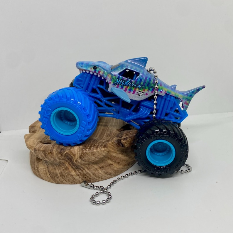 K&ouml;nnte beinhalten: Ein blaues Monstertruck-Spielzeug mit einem Haifisch-Design. Der Truck hat gro&szlig;e blaue Reifen und einen blauen Rahmen. Das Haifisch-Design ist haupts&auml;chlich blau mit bunten Akzenten, und das Wort "Megalodon" ist sichtbar.