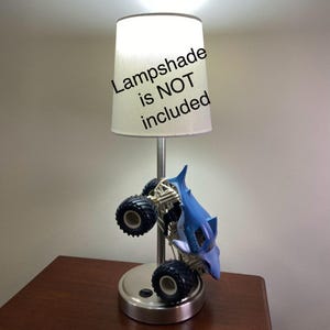 May include: A silver-colored lamp base with a blue monster truck design. The lamp has a white lampshade, with the text "Lampshade is NOT included" on the lampshade.