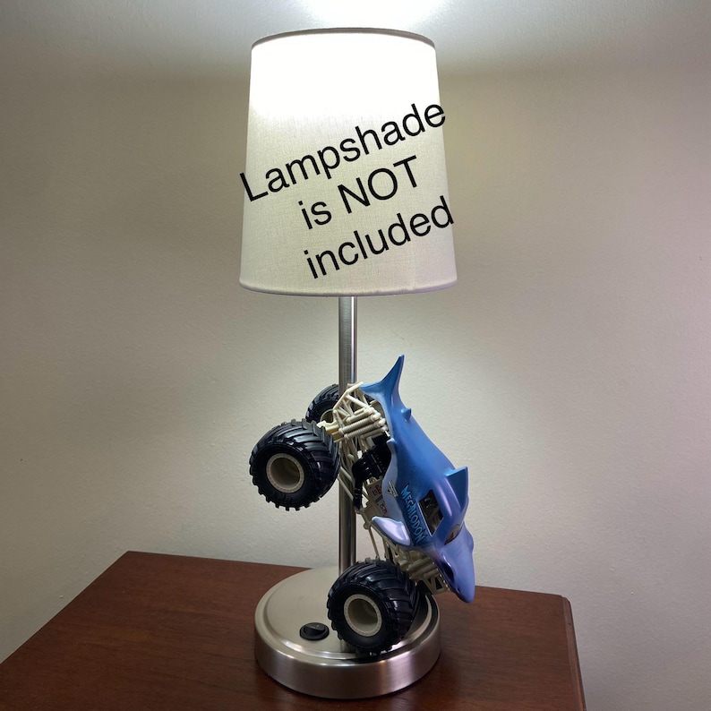 May include: A silver-colored lamp base with a blue monster truck design. The lamp has a white lampshade, with the text "Lampshade is NOT included" on the lampshade.