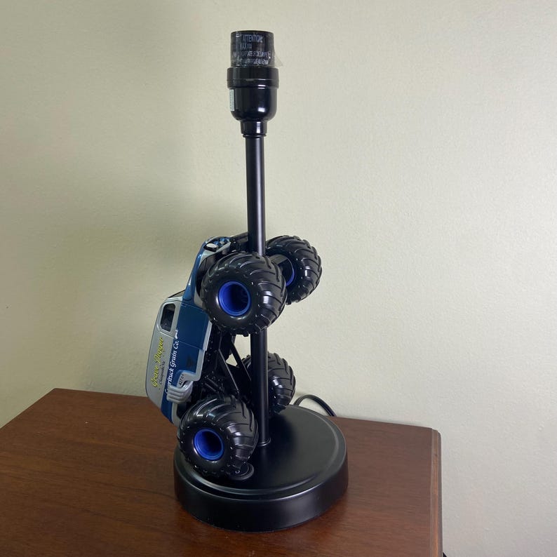 May include: A black table lamp with a monster truck base. The truck is blue and gray with black wheels and blue accents. The lamp has a black pole and a round black base.