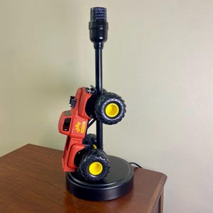 Może przedstawiać: Czerwono-czarna lampa z motywem monster trucka Grave Digger z ż&oacute;łtymi akcentami. Lampa ma czarną podstawę i czarny słup. Opony są czarne z ż&oacute;łtymi środkami. Tekst "Grave Digger" jest widoczny z boku ciężar&oacute;wki.