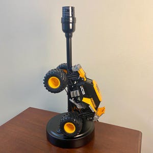 May include: A black table lamp featuring a toy monster truck design. The truck is black and yellow with large, black tires with yellow rims. The lamp has a black base and a tall, black neck.