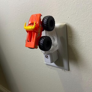 May include: An orange and yellow toy monster truck is plugged into a clear night light. The truck has large black tyres and a yellow bull-like design on the front. The night light is plugged into a white wall socket.