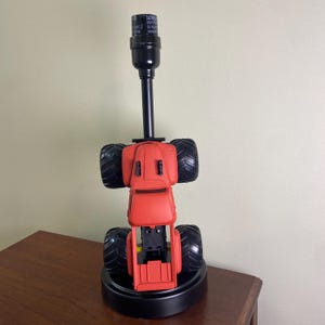 Może przedstawiać: Czerwona lampa w kształcie monster trucka z czarnymi oponami i czarnym słupkiem lampy. Podstawa lampy jest czarna i ma etykietę ostrzegawczą. Lampa stoi na drewnianej powierzchni.