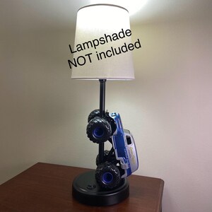 May include: A novelty lamp featuring a blue and silver monster truck design. The truck is mounted on a black base with a black pole supporting a lampshade. The text "Lampshade NOT included" is visible.