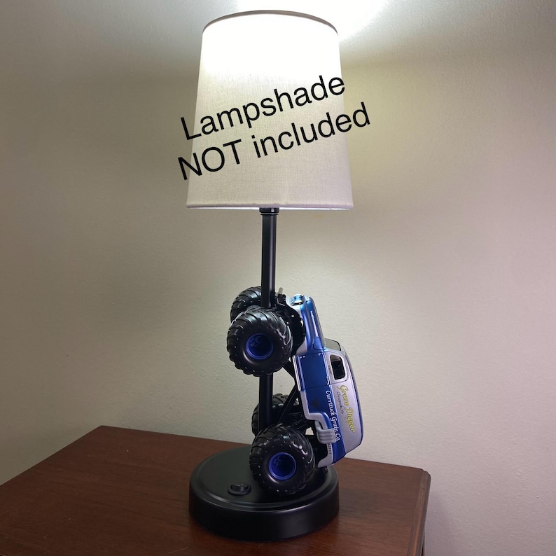 May include: A novelty lamp featuring a blue and silver monster truck design. The truck is mounted on a black base with a black pole supporting a lampshade. The text "Lampshade NOT included" is visible.
