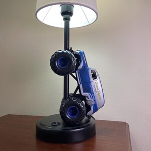 May include: A black table lamp featuring a blue and silver monster truck design. The truck is mounted on a black pole with a gray lampshade. The truck has the text "Grave Digger" and "Currituck Garage Co."