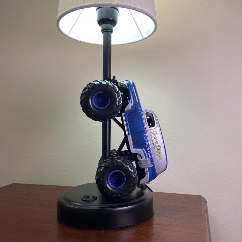 May include: A black table lamp featuring a blue and silver monster truck design. The truck is mounted on a black pole with a gray lampshade. The truck has the text "Grave Digger" and "Currituck Garage Co."
