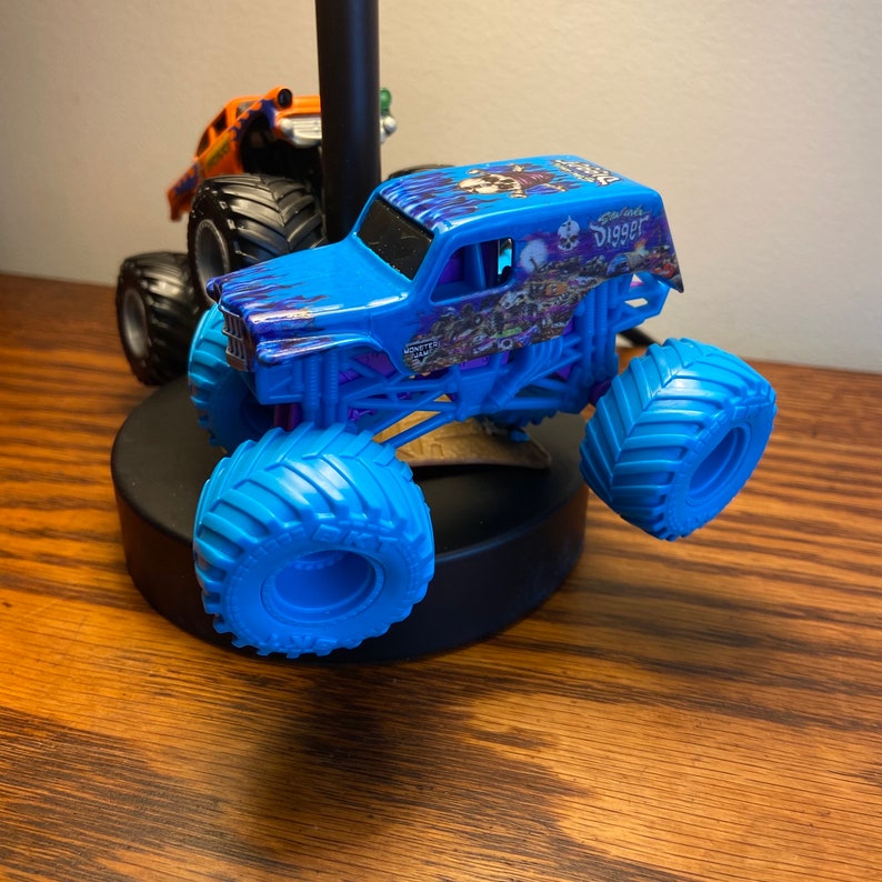 Op de afbeelding: Een blauw monstertruck speelgoed met een schedel en gekruiste beenderen ontwerp aan de zijkant. De truck heeft de woorden "Grave Digger" aan de zijkant gedrukt. De truck staat op een zwarte basis.