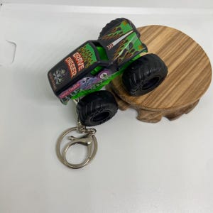 以下が含まれることがあります： 黒と緑の Grave Digger モンスター トラックのおもちゃ。大きな黒いタイヤが付いています。おもちゃのトラックの側面には「Grave Digger」の文字が印刷されています。シルバーのキーリングがトラックに取り付けられています。