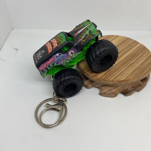以下が含まれることがあります： Grave Diggerのモンスタートラックキーホルダー。おもちゃのトラックは黒と緑で、大きな黒いタイヤが付いています。「Grave Digger」の文字が側面に印刷されています。キーホルダーにはシルバーの留め金とリングが付いています。