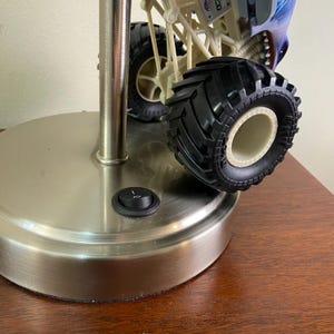 May include: A silver-toned metal lamp base with a black on/off switch. A toy monster truck with black tires and a blue and white body rests on the lamp. The lamp is on a wooden surface.