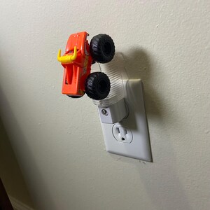 May include: Orange and yellow monster truck night light plugged into a white wall socket. The toy truck has large black tyres and a bull-like design on the front. The night light has a clear plastic cover.