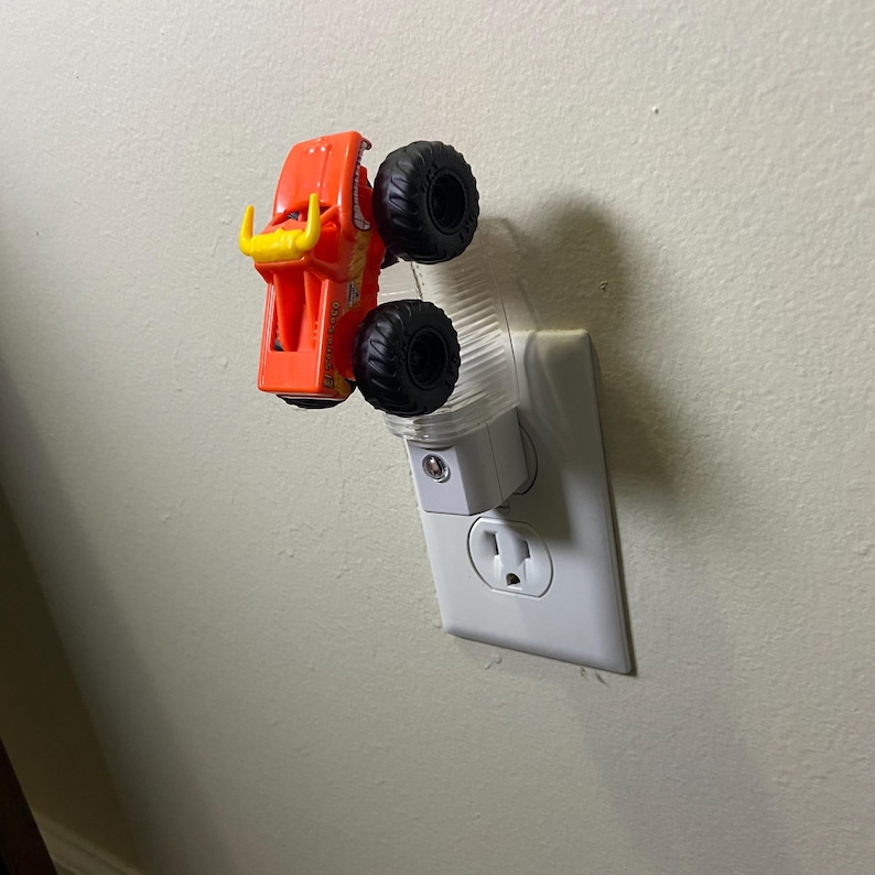 May include: Orange and yellow monster truck night light plugged into a white wall socket. The toy truck has large black tyres and a bull-like design on the front. The night light has a clear plastic cover.