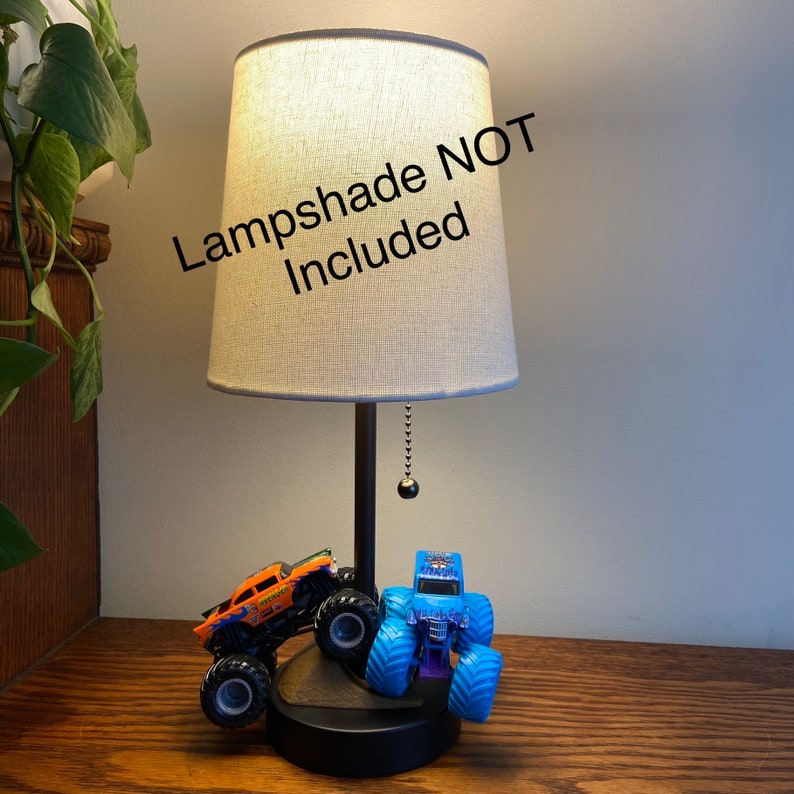 Op de afbeelding: Een zwarte tafellamp met een witte lampenkap van stof. De lampenkap is niet inbegrepen. De lampvoet heeft twee speelgoedmonstertrucks eraan bevestigd. De ene truck is oranje en de andere is blauw.