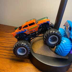 Op de afbeelding: Een oranje en groene monstertruck speelgoed met het woord "Avenger" aan de zijkant. De truck heeft grote zwarte wielen en staat op een bruin oppervlak.